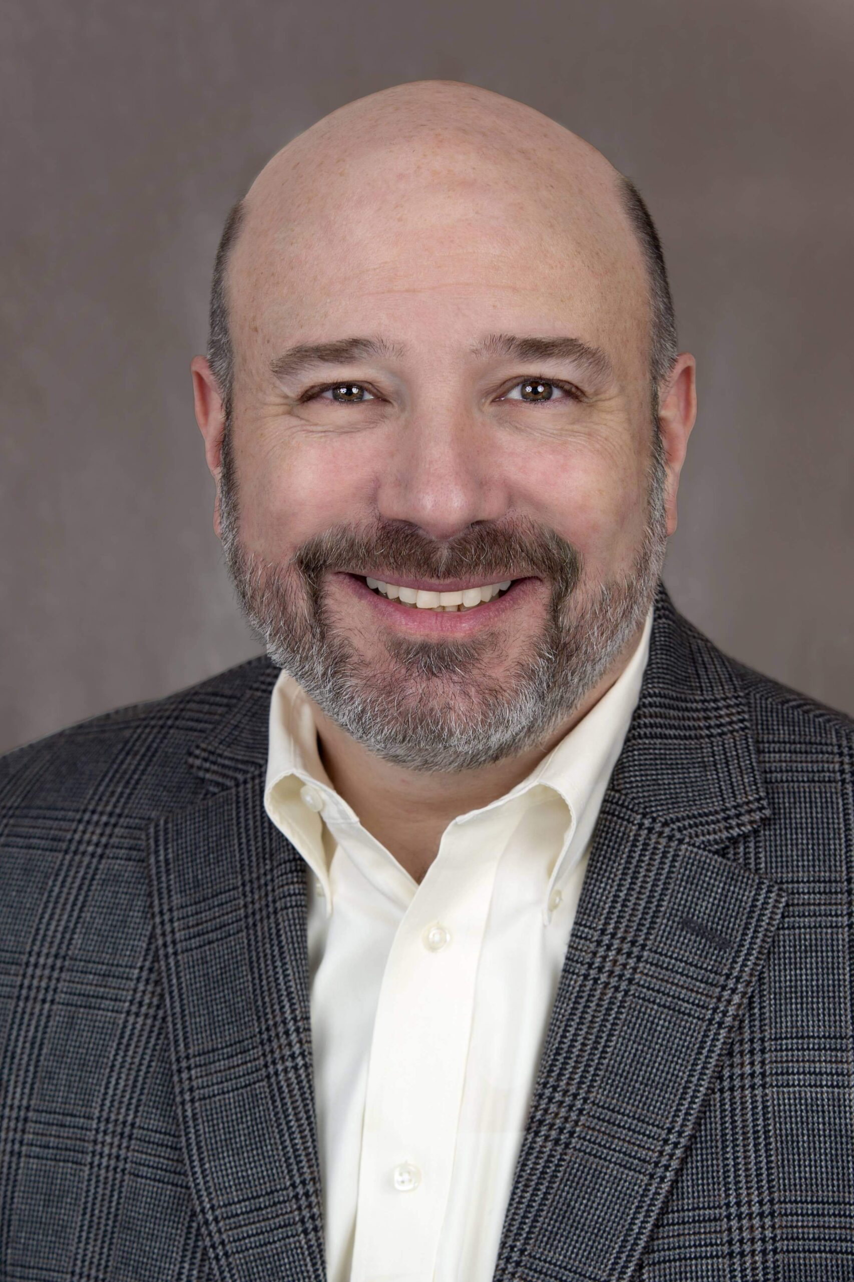 A bald man with a gray beard and mustache, who could easily be mistaken for Eric R. Posmantier, is smiling. He is wearing a gray checkered suit jacket over a white dress shirt. The background is a plain, gray backdrop.