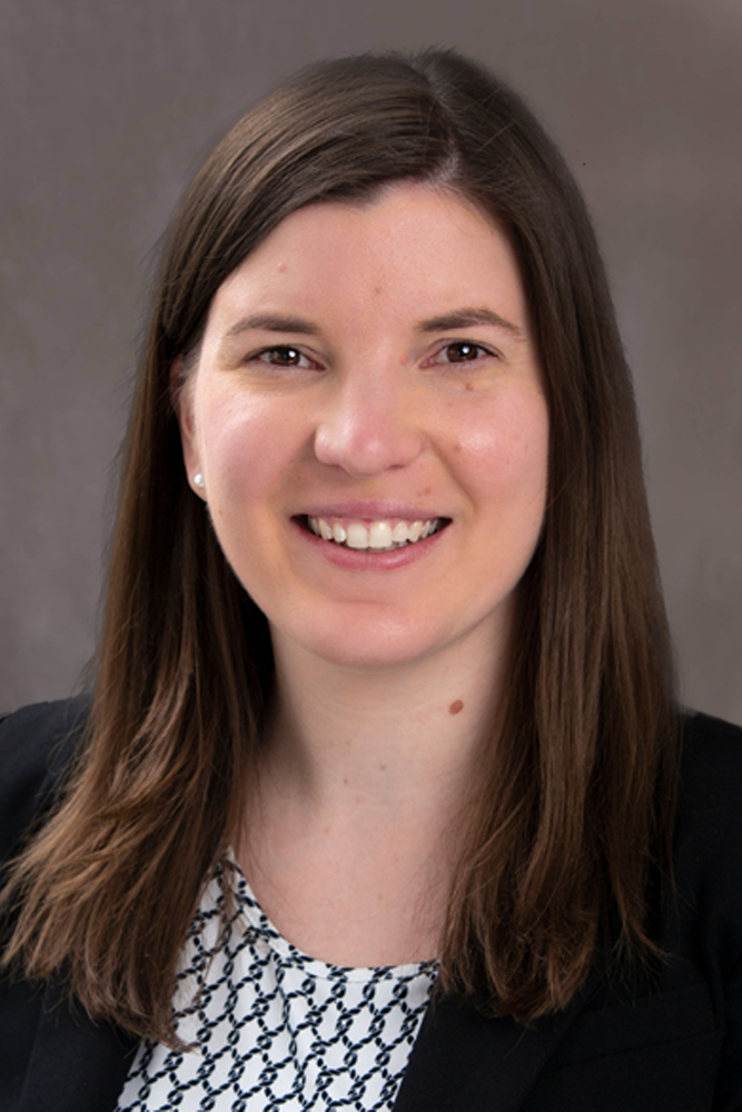 Kimberly A. Stokes, a woman with long, straight brown hair, smiles warmly. She is wearing a black blazer over a patterned blouse. The background is a plain, grey gradient.