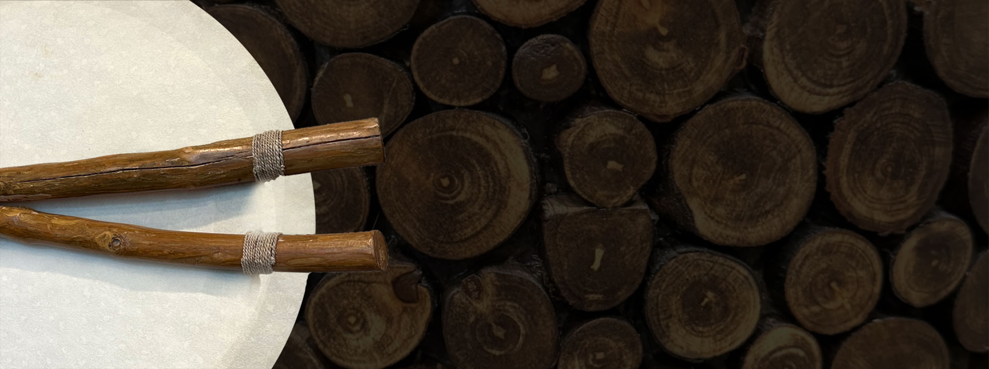 A white drum with wooden drumsticks lies on a surface with a background of stacked, cut wooden logs. The drumsticks have a natural, rugged appearance, and the logs show various sizes and circular patterns.